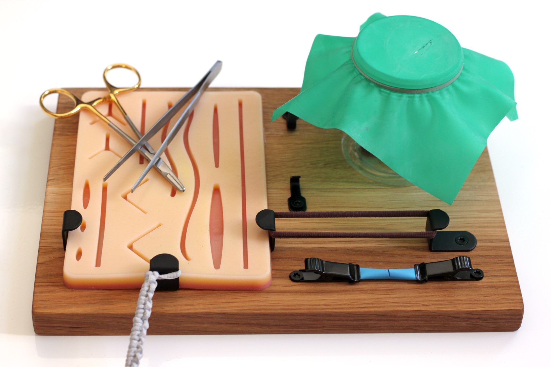 Suture practice kit set up: oak board with silicone pad, needle holder and forceps, a cord on one hook, elastic bands between the hooks, a tube in the clamps and a rubber sheet over the cylinder